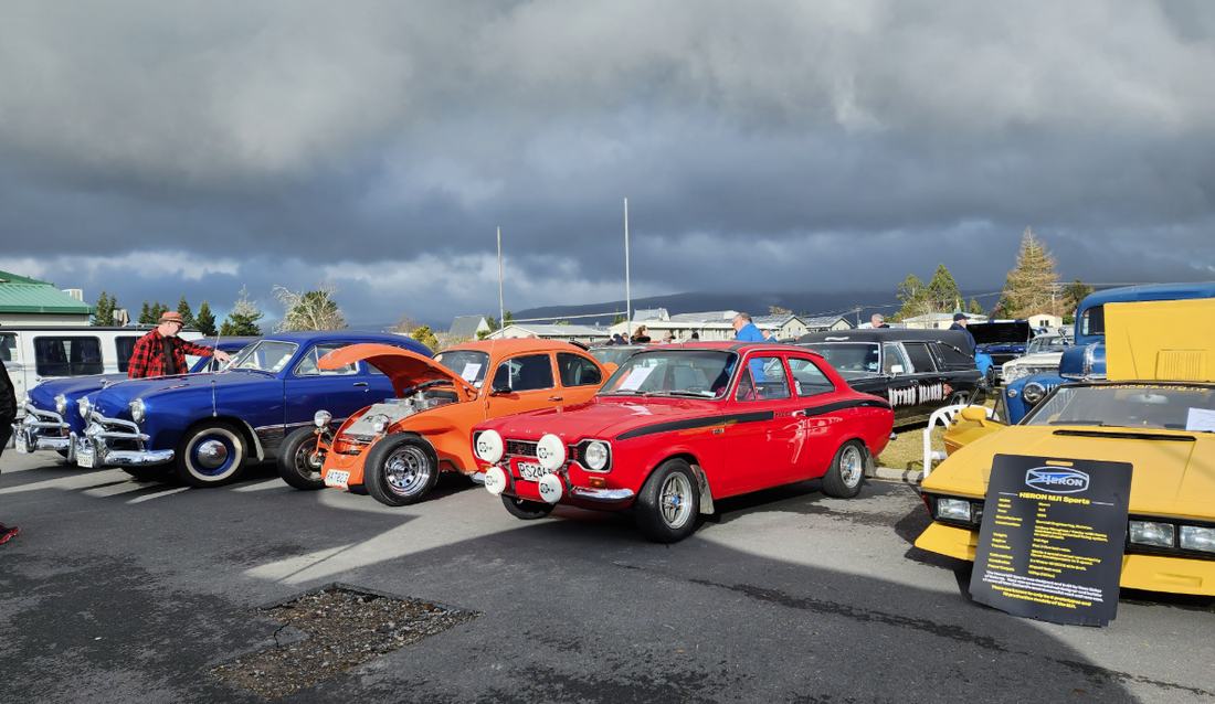 Row of classic cars at a previous show, including a blue 1949 Ford, an orange hot rod and a red Ford Escort Mexico